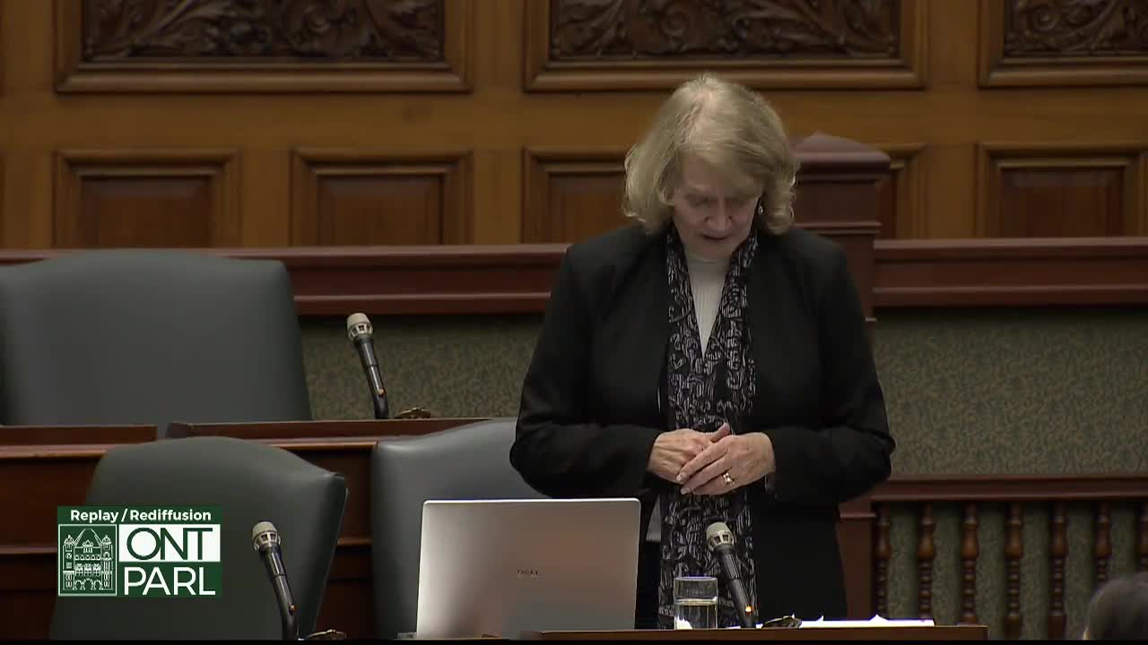 A woman in a black blazer and patterned scarf stands at a podium in the Legislative Assembly of Ontario, speaking with her hands clasped. A laptop sits in front of her, and two microphones are visible.
