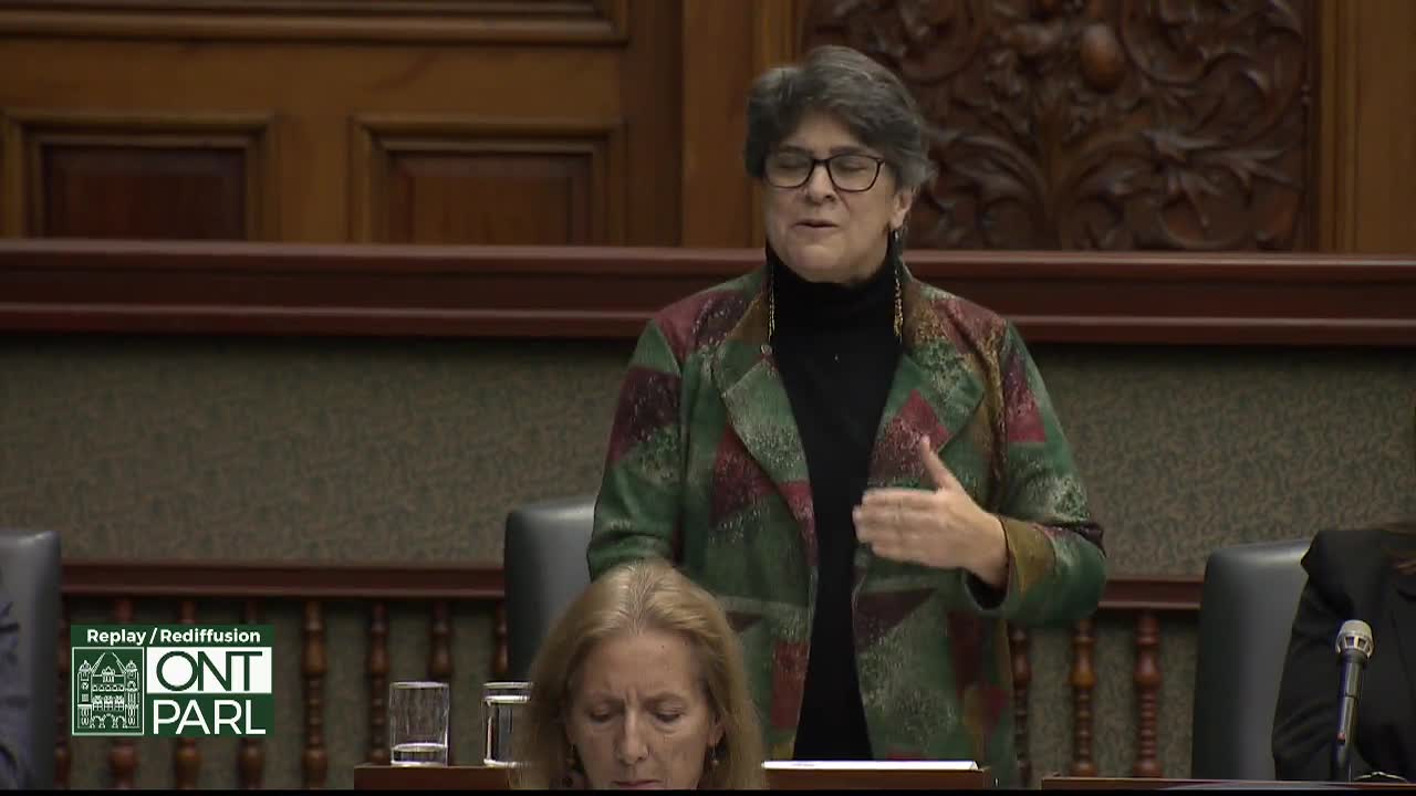 A woman in a patterned jacket gestures with her right hand, speaking at the Legislative Assembly of Ontario. Another woman sits below, looking down, while the replay logo of the Ontario Parliament is visible.
