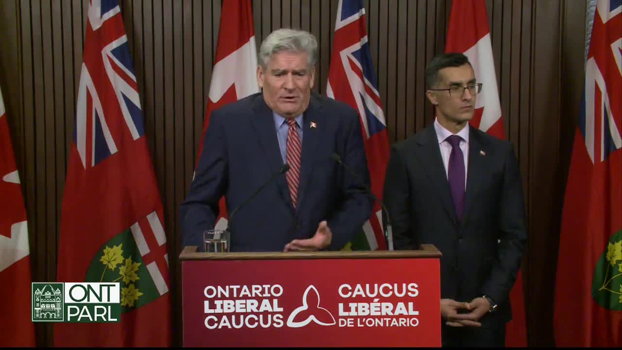 A man in a navy suit gestures emphatically at a podium bearing the Ontario Liberal Caucus logo, flanked by flags of Canada and Ontario. Beside him, another man in a dark suit stands still, watching the speaker.
