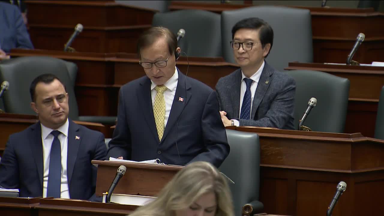 A man in a navy suit and yellow tie is speaking from a podium in the Legislative Assembly of Ontario, reading from a paper. Two other men in suits sit nearby, listening intently.
