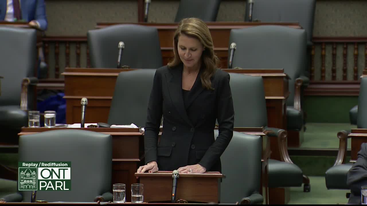 In the Legislative Assembly of Ontario, a woman in a black suit stands at a podium, speaking. The room is quiet, and she appears to be reading from notes.
