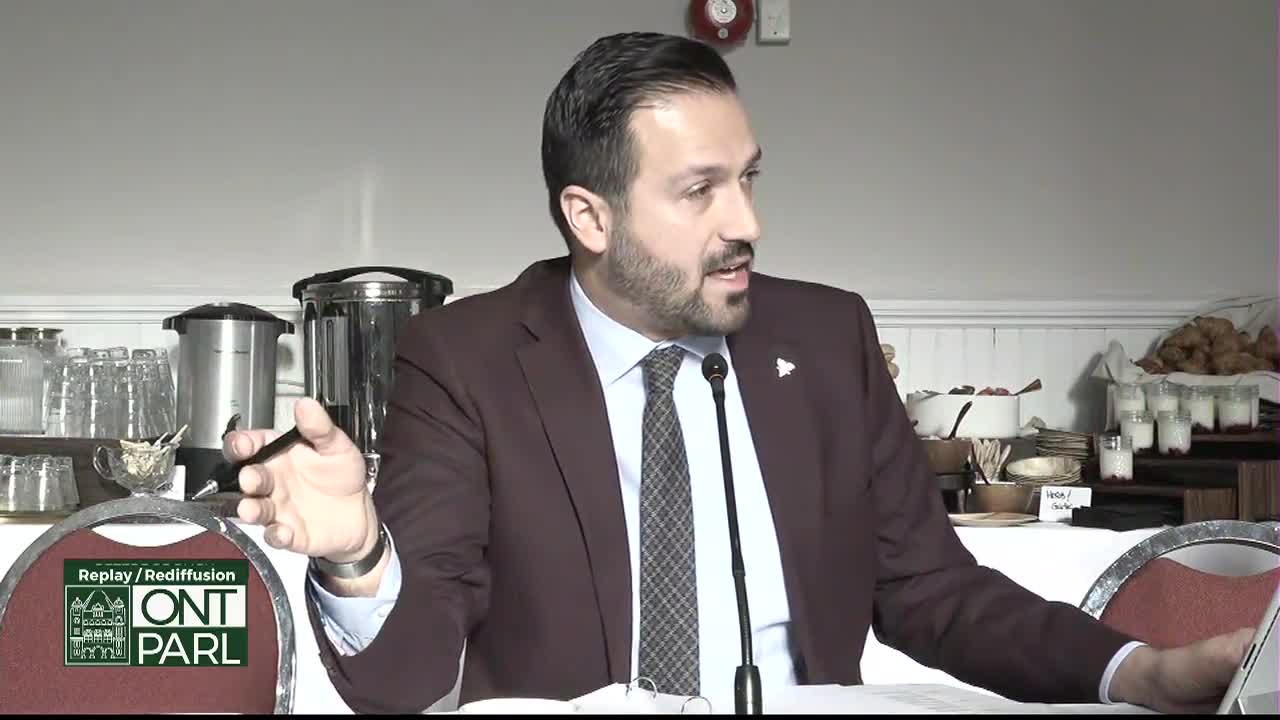 A man in a maroon suit gestures with a pen, speaking into a microphone at a table. Behind him, a buffet is set up, and the Legislative Assembly of Ontario's logo is visible on a nearby chair.
A man in a maroon suit gestures with a pen, speaking into a microphone at a table. Behind him, a buffet is set up, and the Legislative Assembly of Ontario's logo is visible on a nearby chair.