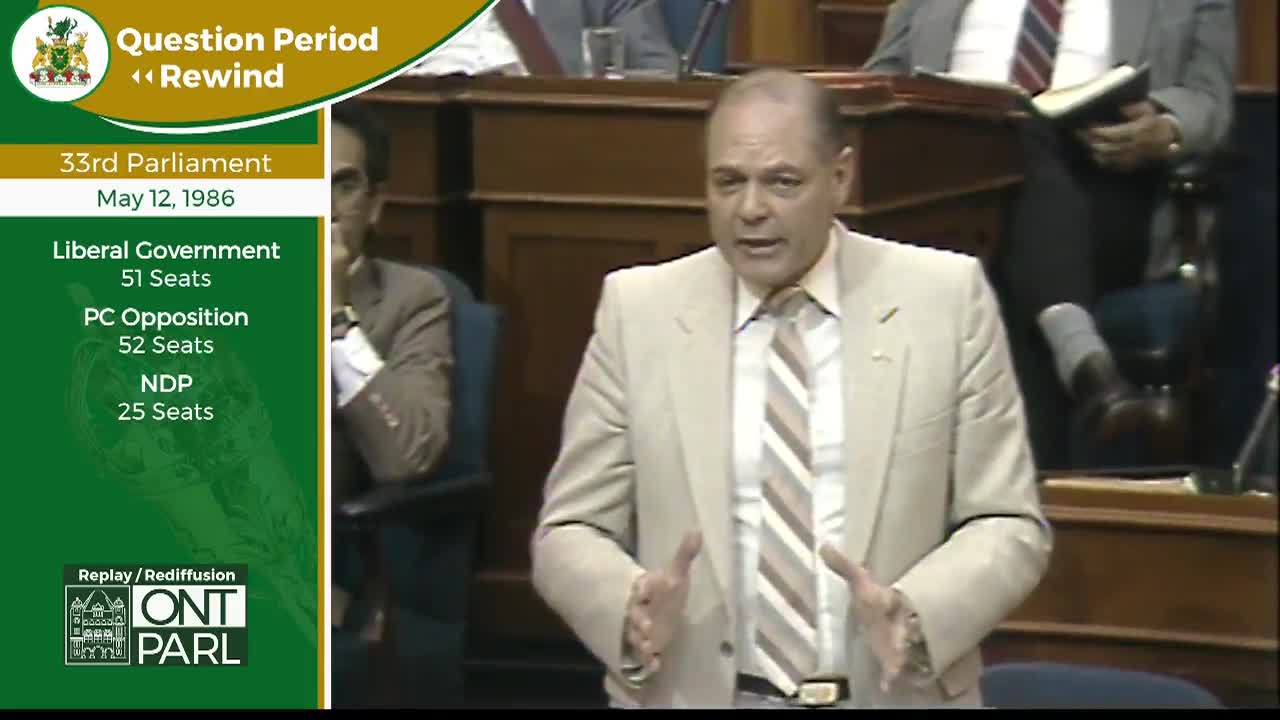 A man in a tan suit speaks with his hands gesturing, while another man in the background holds a book. This is the Legislative Assembly of Ontario during Question Period on May 12, 1986.