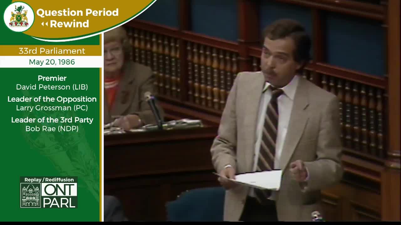 A man in a tan suit stands at a podium, holding a paper and speaking. Behind him, a woman with short blonde hair sits at another desk in the Legislative Assembly of Ontario.