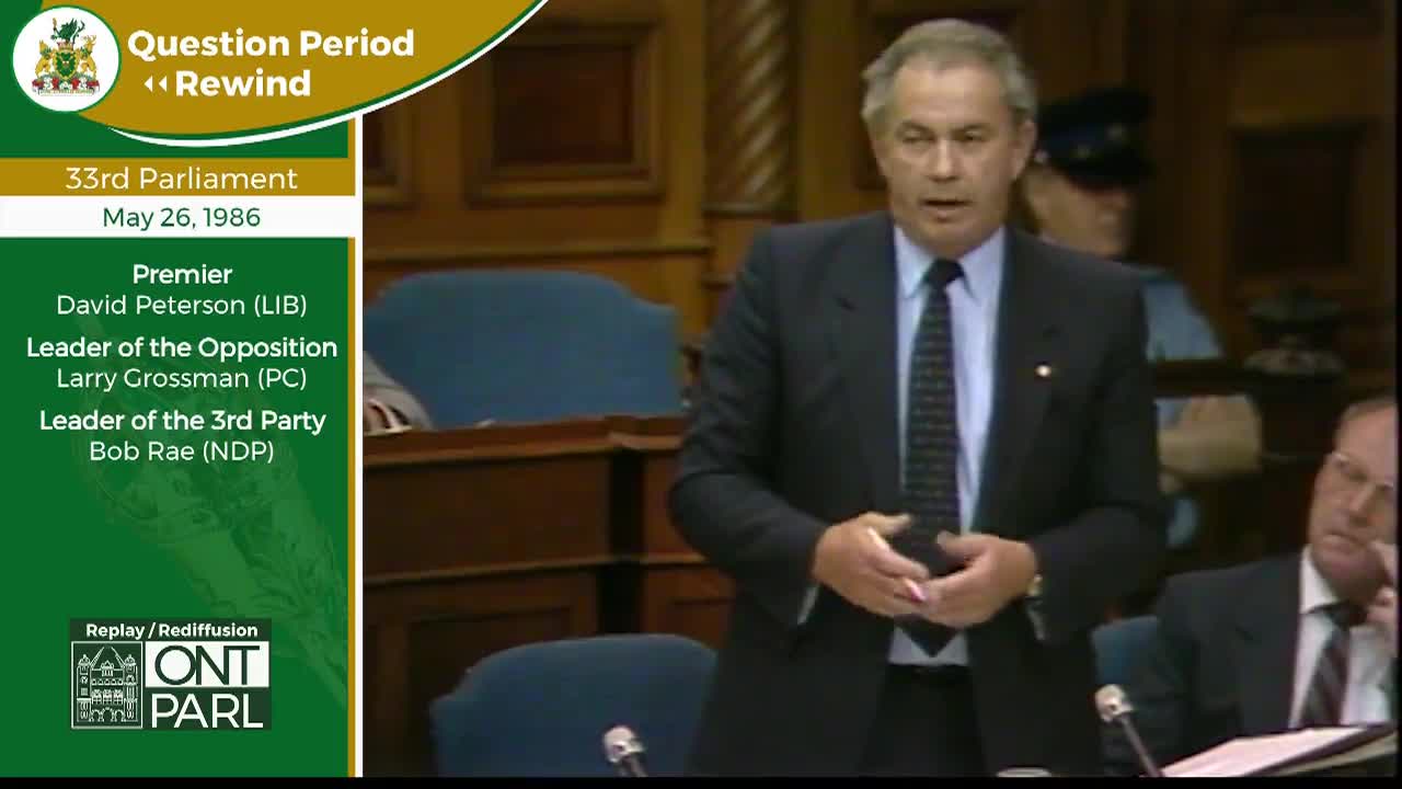 A man in a dark suit and tie stands speaking, his hands clasped loosely in front of him. Behind him, a person in a dark uniform stands at attention.