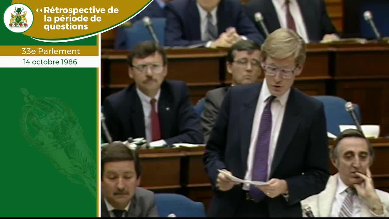 A man in a dark suit and purple tie stands at a podium, holding a paper. He speaks to the assembly while another man in a white jacket leans forward, listening intently. A man in a dark suit and purple tie stands at a podium, holding a paper. He speaks to the assembly while another man in a white jacket leans forward, listening intently.