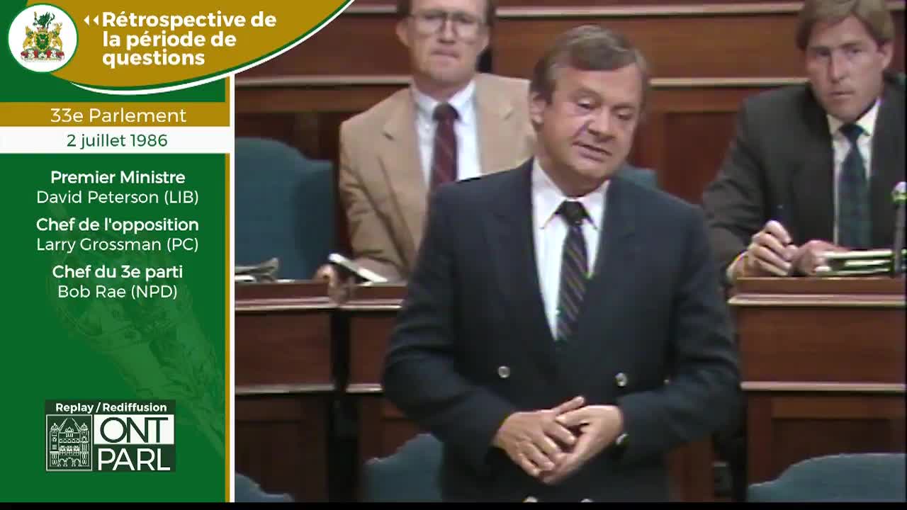 A man in a dark suit stands at a podium, his hands clasped. Behind him, another man in a tan jacket and glasses watches intently. This is the Legislative Assembly of Ontario, during a question period in July 1986.