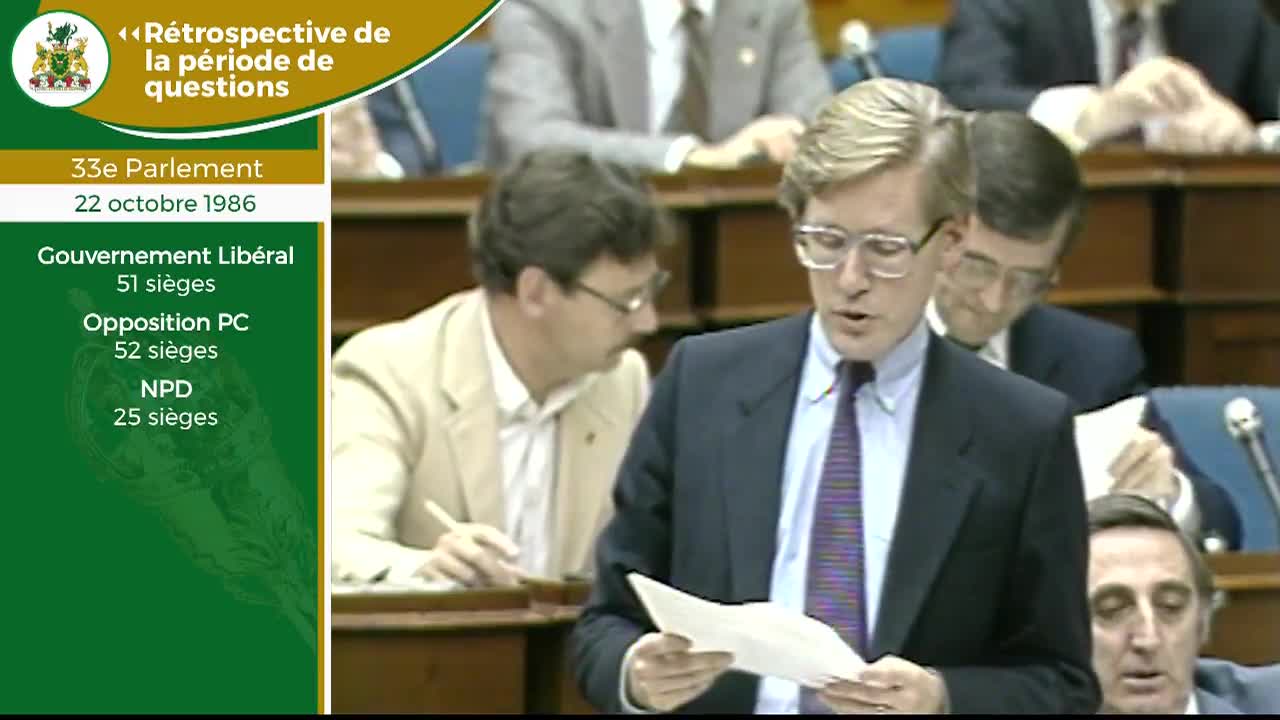 A man in a dark suit and tie stands at a podium, holding a paper. He speaks while others in the Legislative Assembly of Ontario listen.