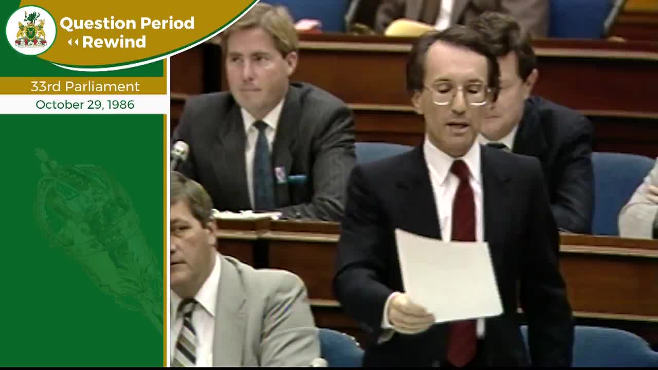 A man in a dark suit and red tie stands, holding a white paper. He speaks to the room, facing the benches where other men in suits sit. The date October 29, 1986, appears on a green banner beside the scene.