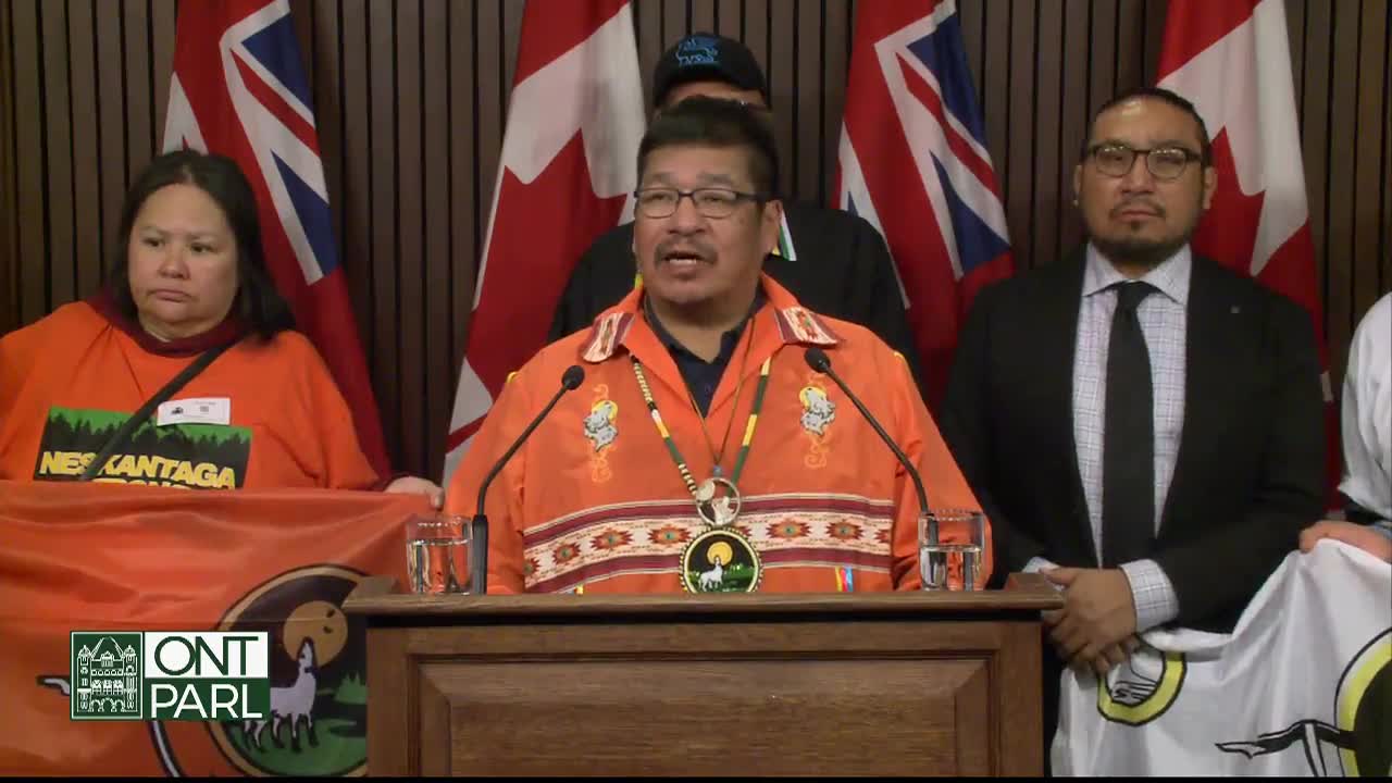 A man in an orange shirt speaks at a podium, flanked by people holding flags. Behind them, the flags of Ontario and Canada hang.
