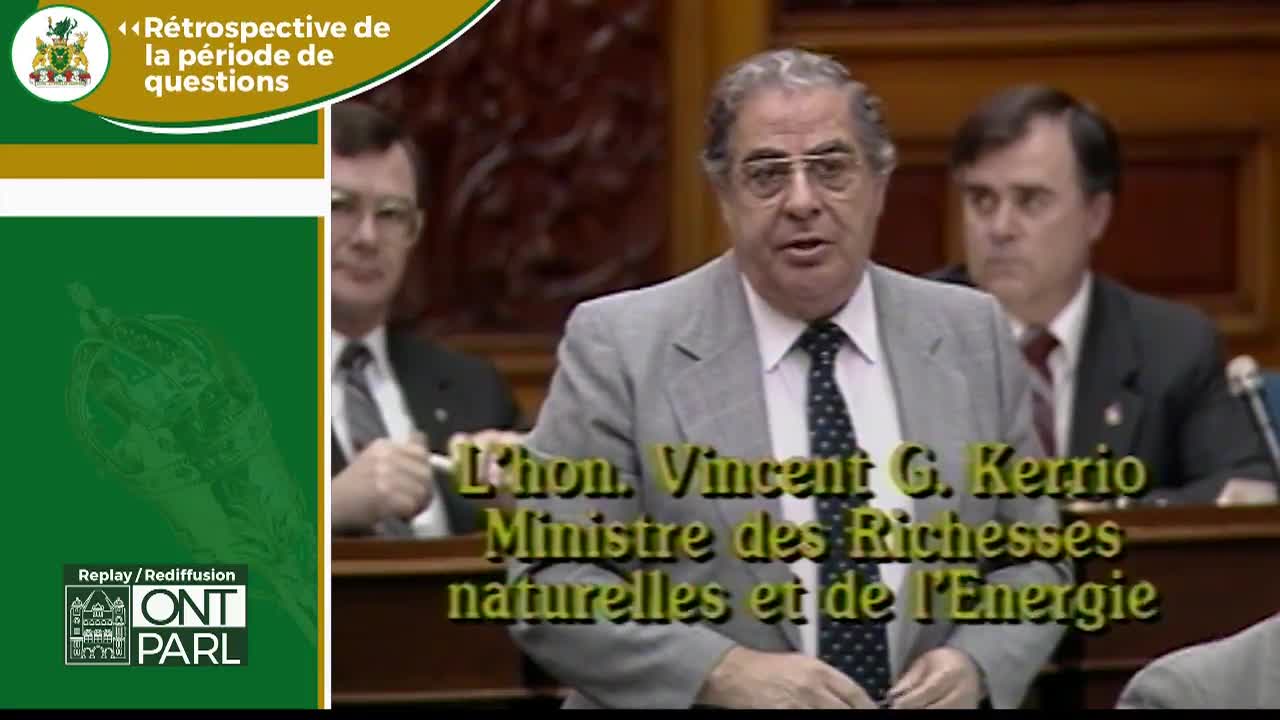 The Honourable Vincent G. Kerrio, Minister of Natural Resources and Energy, speaks at the Legislative Assembly of Ontario. He gestures with his right hand while another man in a suit and tie looks on from behind.