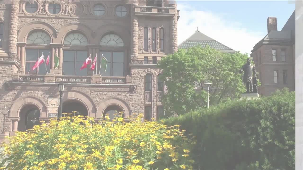 Flags snap in the breeze above the Legislative Assembly of Ontario. A bronze statue stands tall against the building's stone facade.