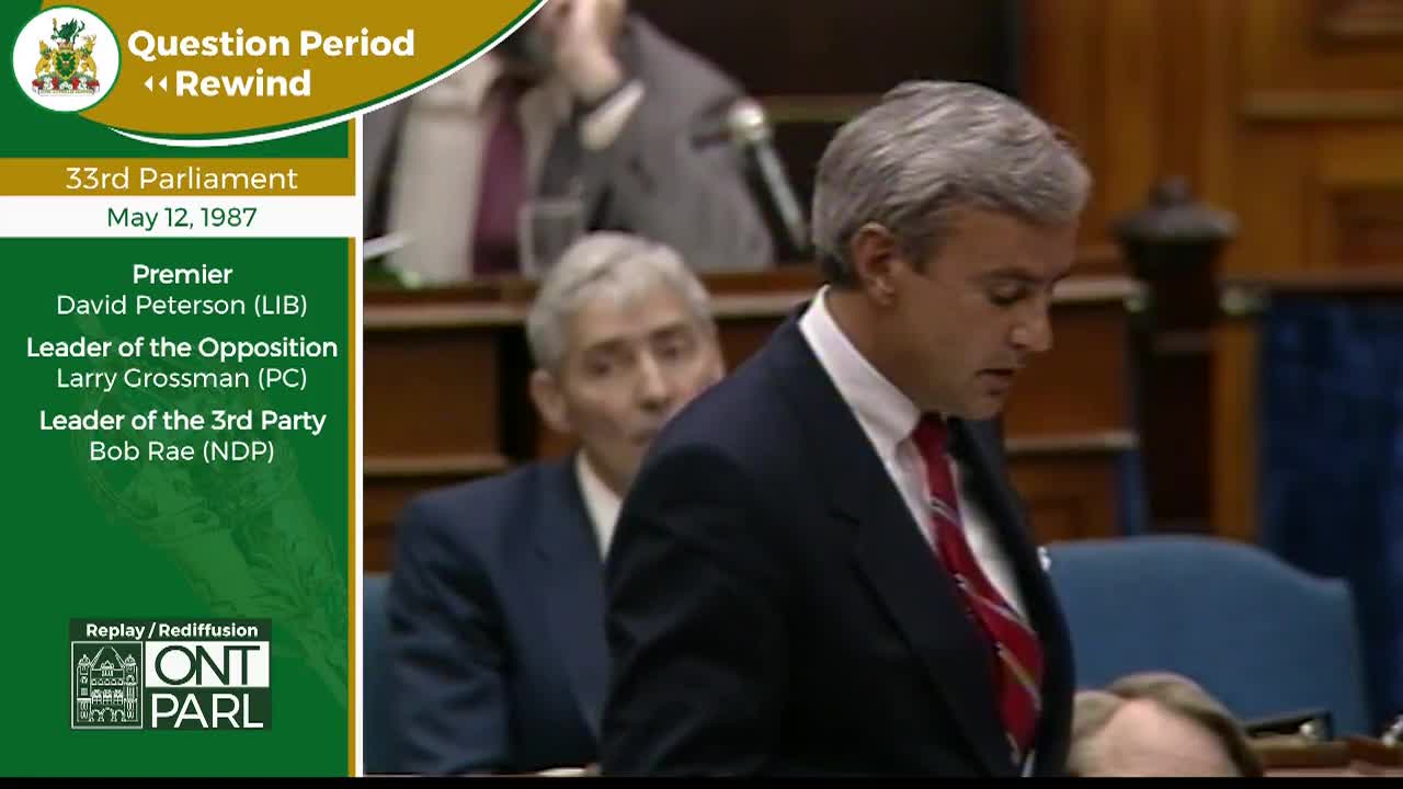 A man in a dark suit and red tie speaks, his head bowed slightly. Behind him, another man in a suit looks towards the speaker. The scene unfolds during Question Period in the Legislative Assembly of Ontario on May 12, 1987.