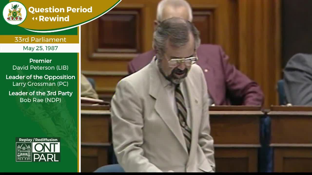 A man in a light-colored suit stands at a podium in the Legislative Assembly of Ontario, speaking with his head bowed. Behind him, other members of parliament are seated in rows of wooden benches.