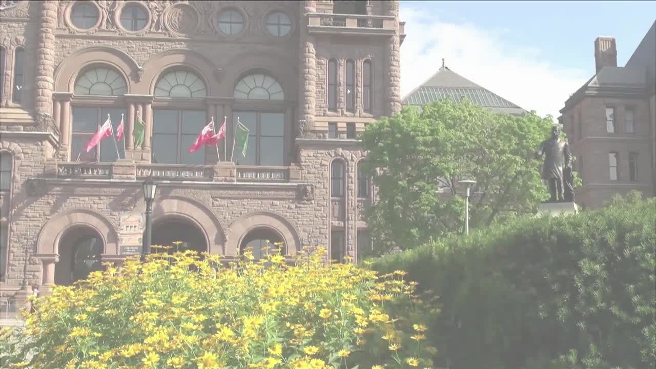 Flags snap in the breeze atop the Legislative Assembly of Ontario. A bronze statue stands stoically amidst the greenery.