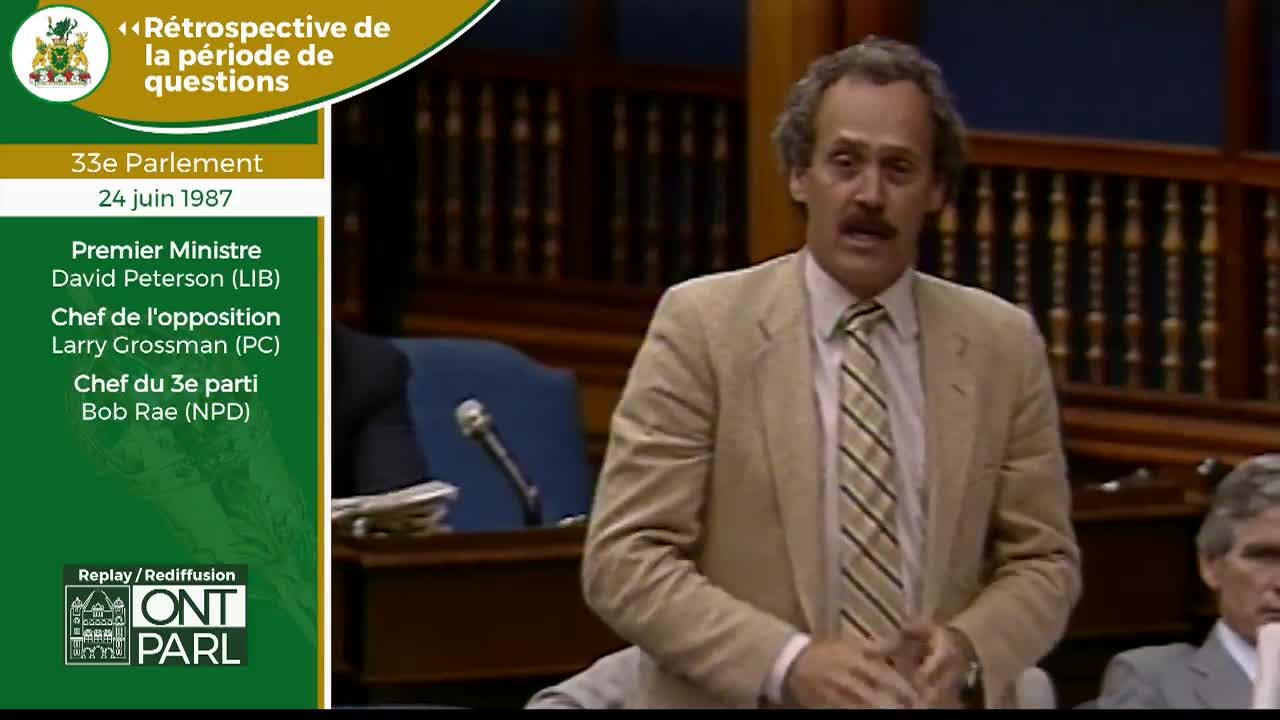 A man in a tan suit and striped tie speaks animatedly in the Ontario Legislative Assembly. He gestures with his hands as he addresses the floor.