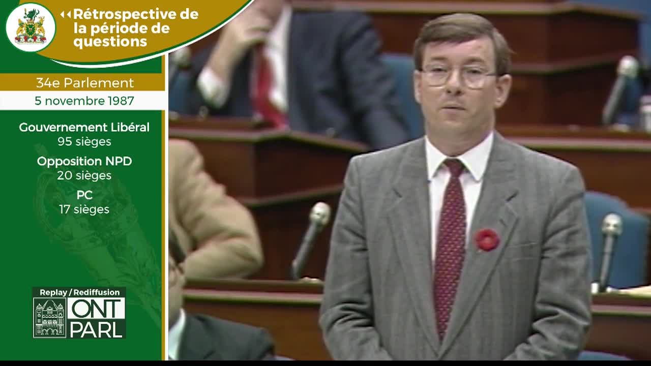 A man in a grey suit stands at a podium, speaking. He wears a red poppy on his lapel. Behind him, other figures are seated in tiered rows, their faces indistinct.