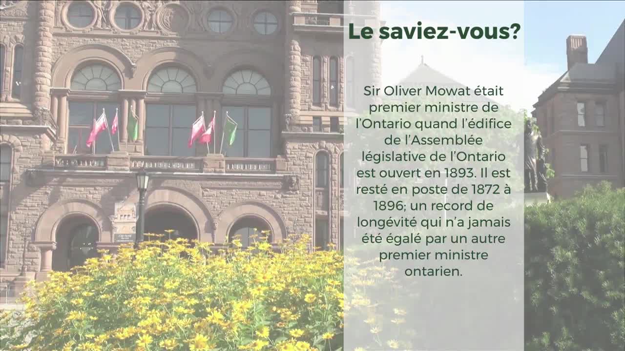 Flags flutter on the balcony of the Legislative Assembly of Ontario. A bright yellow flower bed blooms in front of the historic stone building.