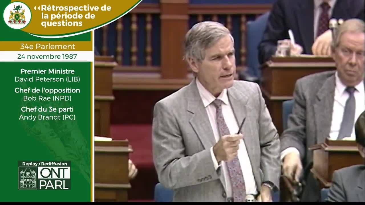 The Premier speaks, gesturing with a pen. Beside him, another man listens intently.