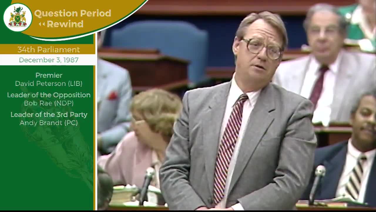 A man in a grey suit and striped tie speaks from his seat in the Legislative Assembly of Ontario. Behind him, other members of parliament listen, some with their heads tilted.