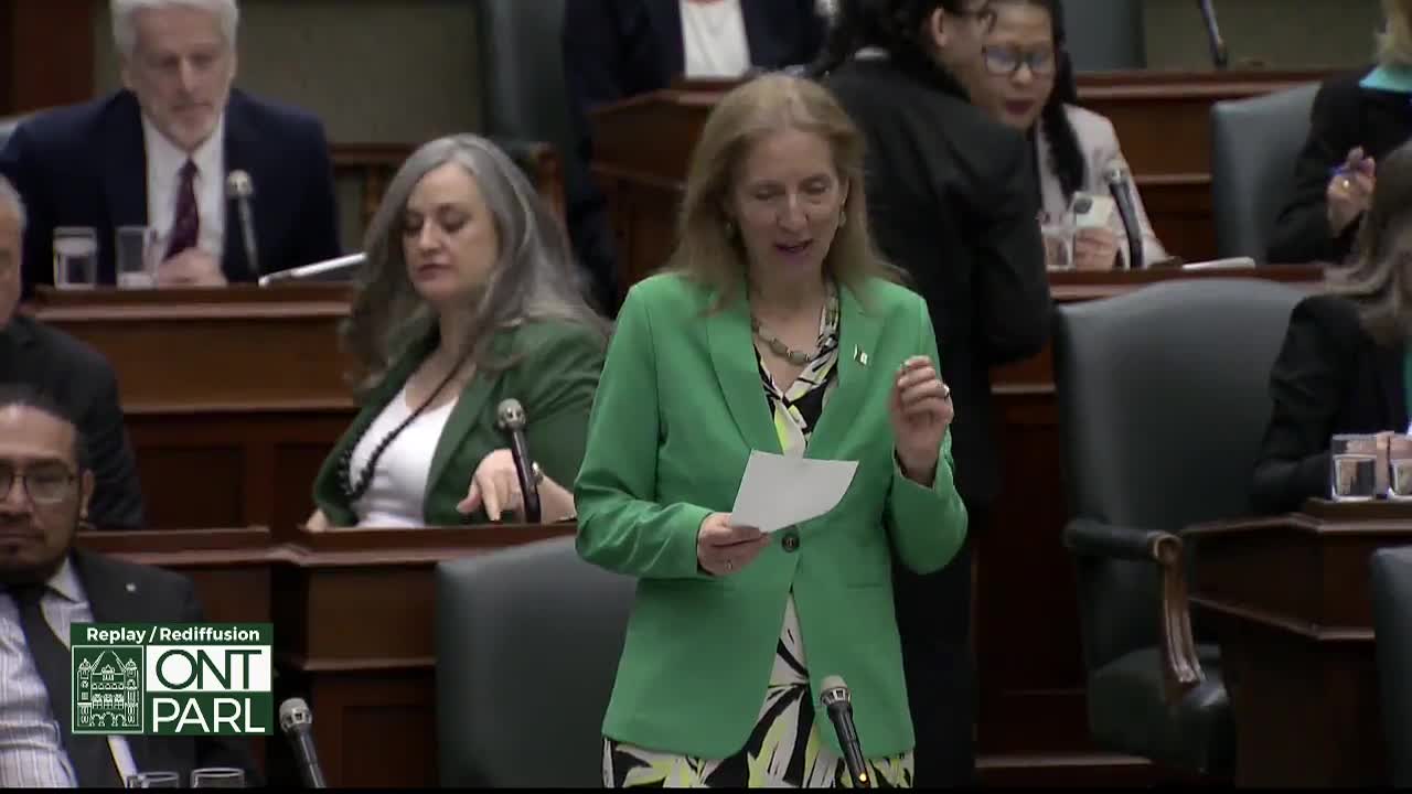 A woman in a bright green jacket stands at a podium, holding a piece of paper. She gestures with her right hand as she speaks to the assembled members of the Legislative Assembly of Ontario.