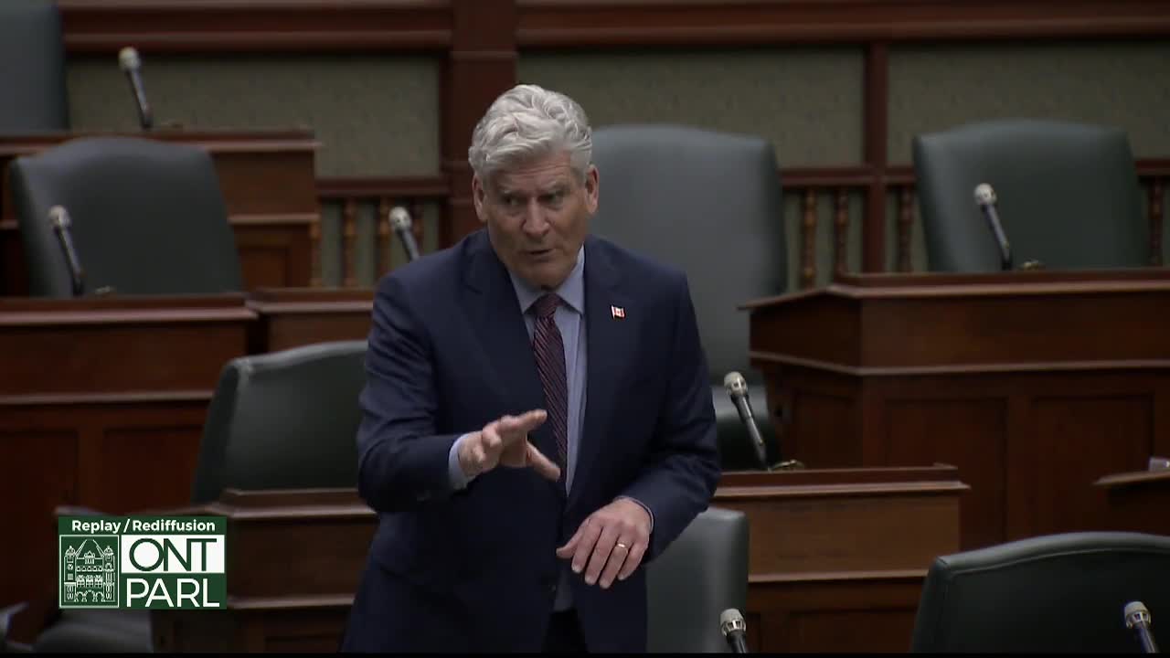 A man in a dark suit gestures with his hands as he speaks in the Legislative Assembly of Ontario. Rows of empty seats and microphones fill the background.