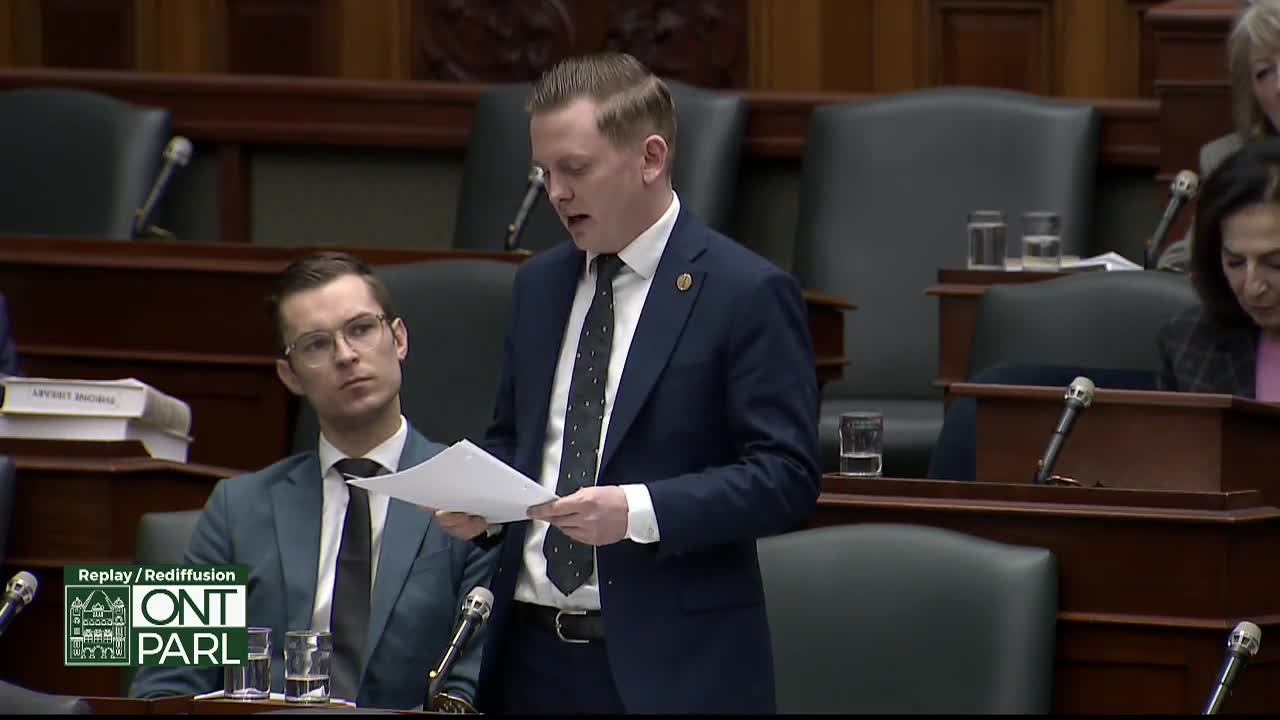 A man in a dark suit stands at a podium, reading from a document. Beside him, another man in a suit and glasses listens intently. They are in the Legislative Assembly of Ontario.