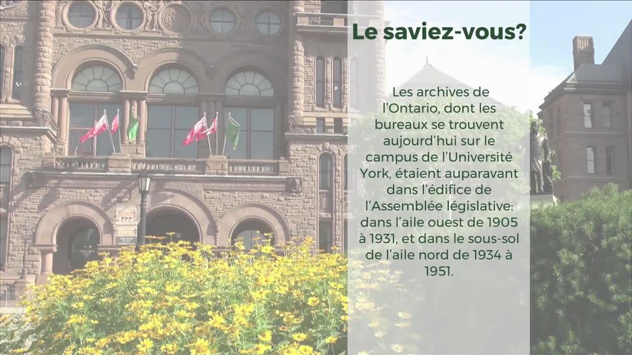 Flags flutter on the Legislative Assembly building. Yellow flowers bloom in front of the stone structure.