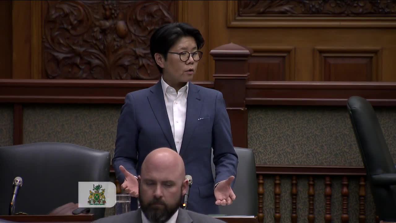 A woman in a blue suit stands at a podium, speaking with her hands gesturing. The Ontario Legislative Assembly's distinctive wood paneling surrounds her. 
