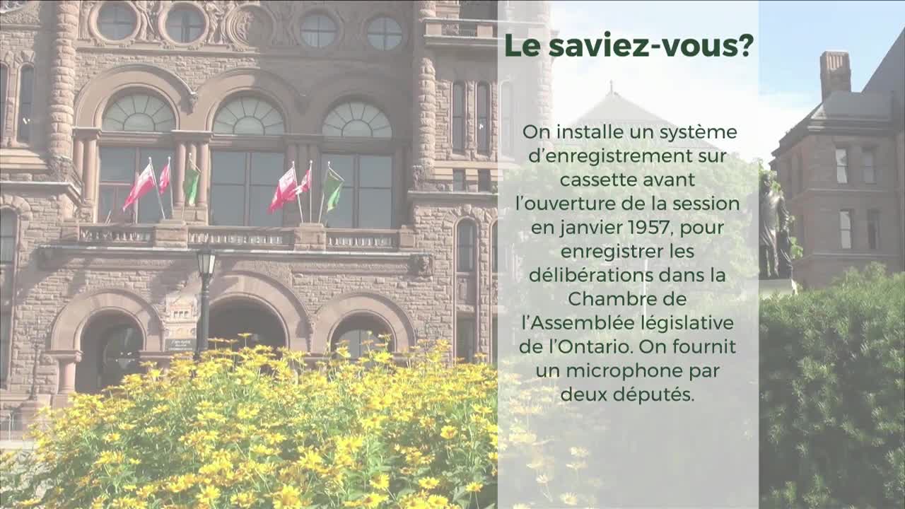 Flags flutter on the balcony of the Legislative Assembly of Ontario. A bright yellow floral display blooms in front of the stone building.