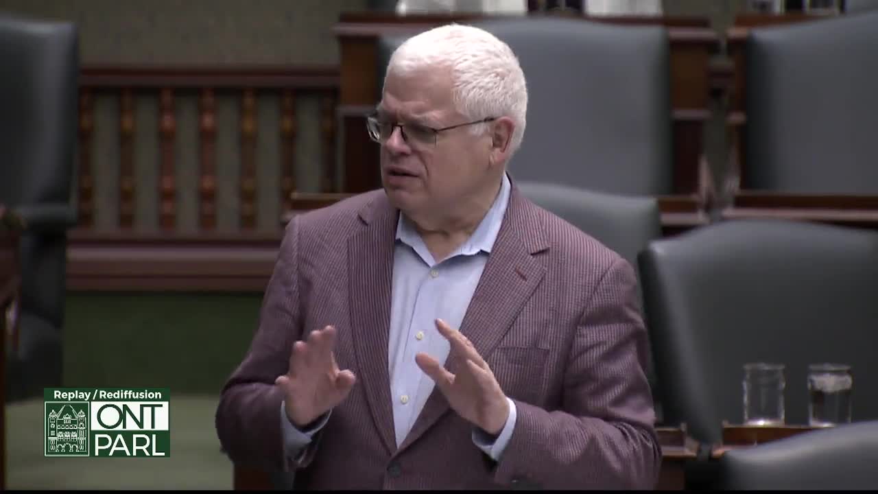 A man in a pinstripe jacket gestures with both hands while speaking in the Legislative Assembly of Ontario. He wears glasses and a light blue shirt.