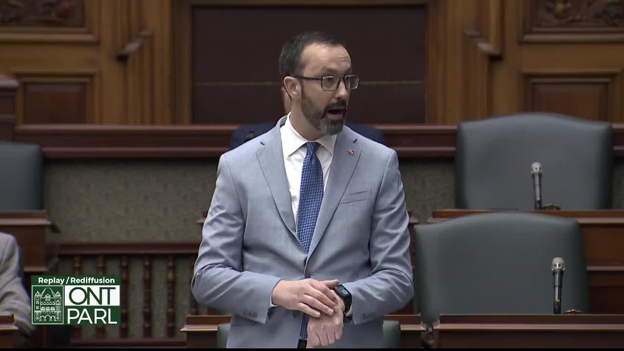 A man in a light blue suit stands at a podium, speaking. He gestures with his left hand, glancing at his watch. The wood-paneled chamber of the Legislative Assembly of Ontario surrounds him.