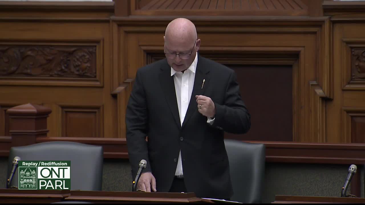 A man in a black suit stands at a podium in the Legislative Assembly of Ontario. He adjusts his tie as he speaks, the ornate wood paneling of the chamber behind him.