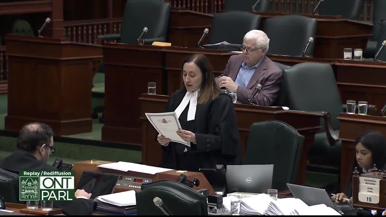 A woman in a black robe and white collar stands at a podium, reading from a document. Behind her, a man in a purple jacket leans back in his seat.