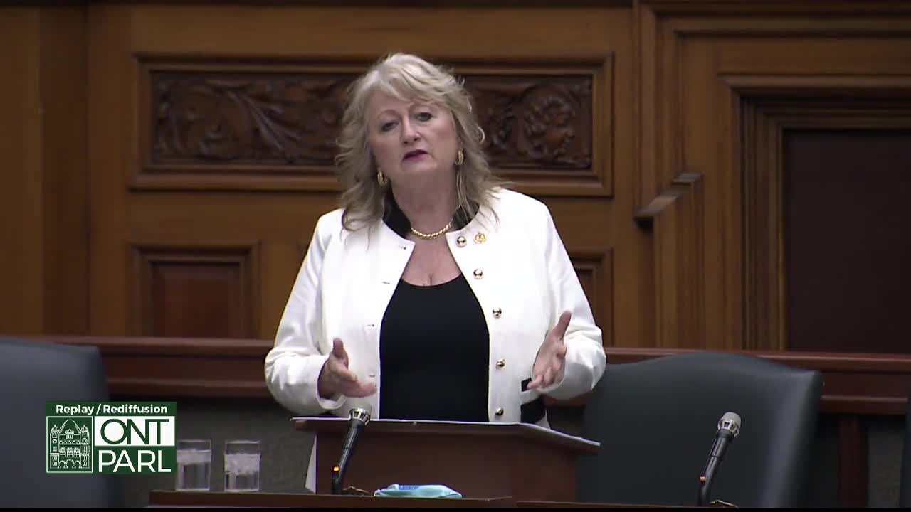 A woman in a white jacket speaks with her hands gesturing. She stands at a podium in what appears to be the Legislative Assembly of Ontario.