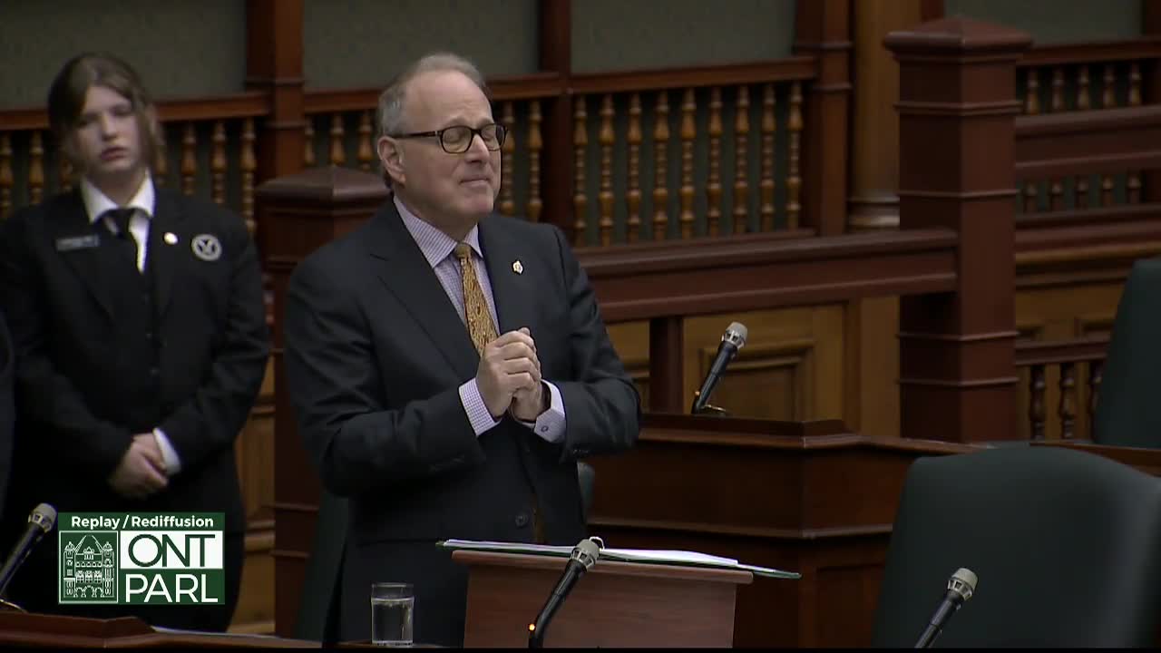 A man in a dark suit clasps his hands together in front of a wooden podium. To his left, a young person in a black uniform stands attentively.
