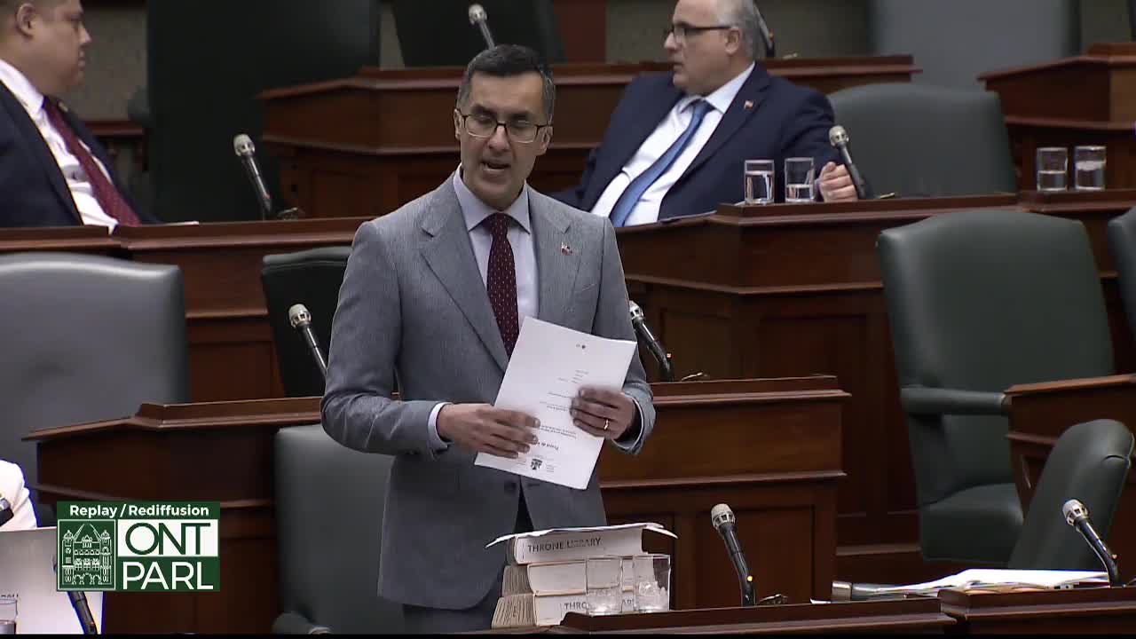 A man in a grey suit stands at a podium, holding papers. He speaks while another man in a blue suit watches from behind.