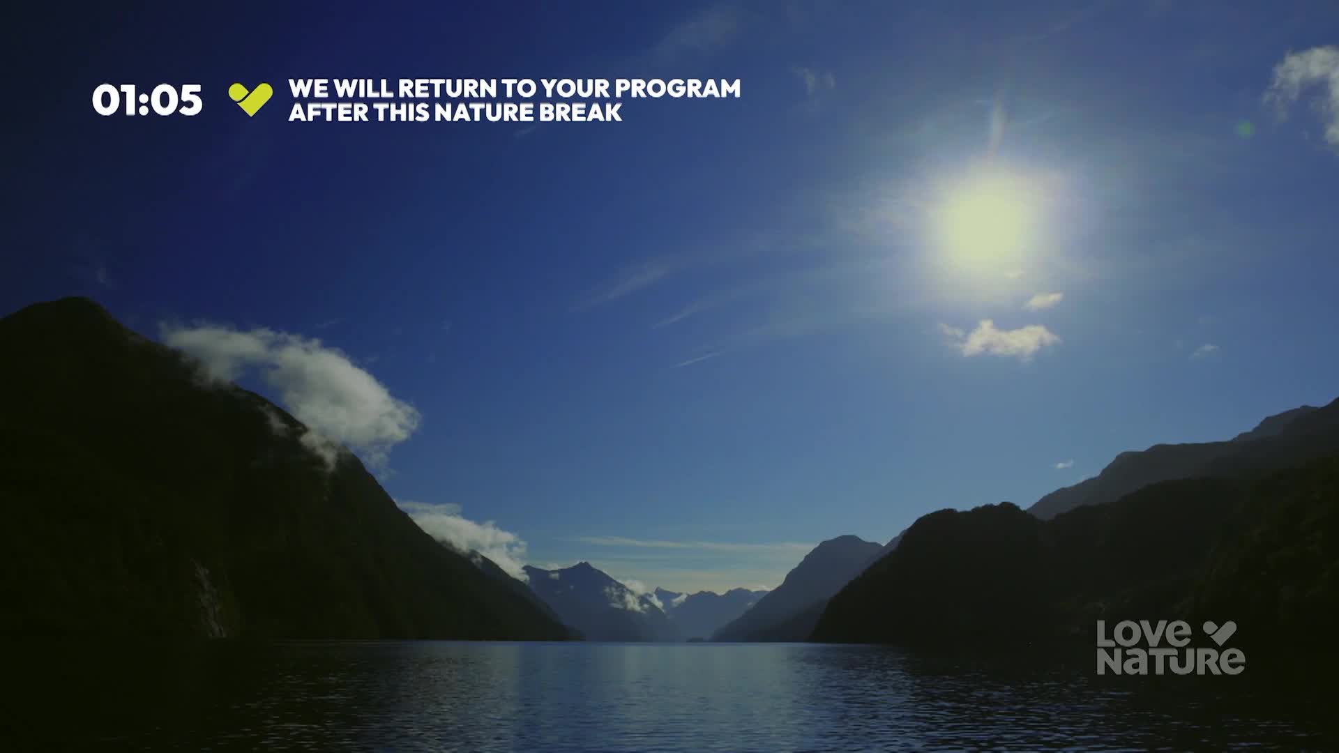 The sun blazes above a calm lake, its light reflecting off the water. Dark mountains frame the scene, with wisps of clouds clinging to their peaks.
