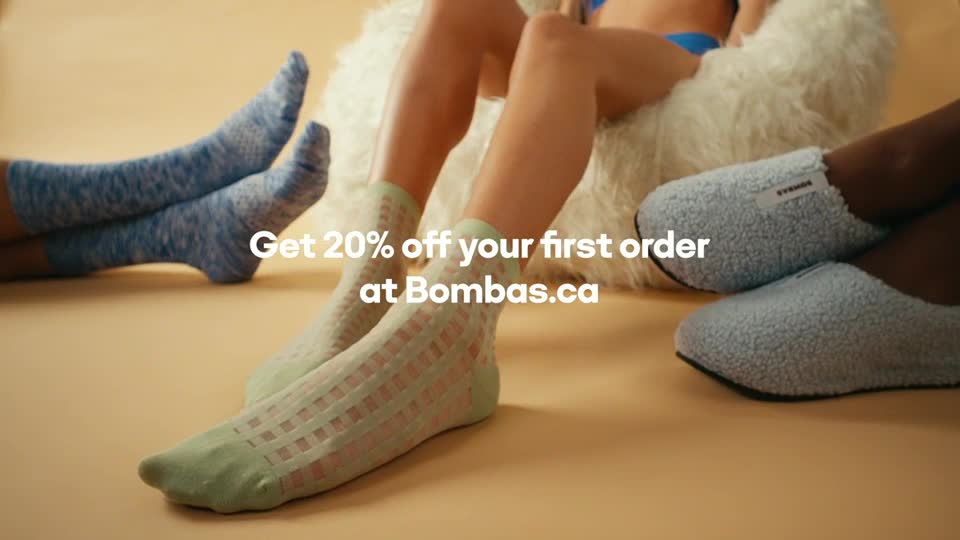 Several pairs of legs, each wearing different socks or slippers, are casually arranged on a pale surface. The text "Get 20% off your first order at Bombas.ca" is overlaid on the scene.
