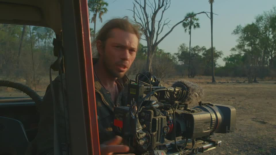 A man in a vehicle is holding a large camera, looking directly at me. The camera's lens is pointed towards the dry landscape outside.
