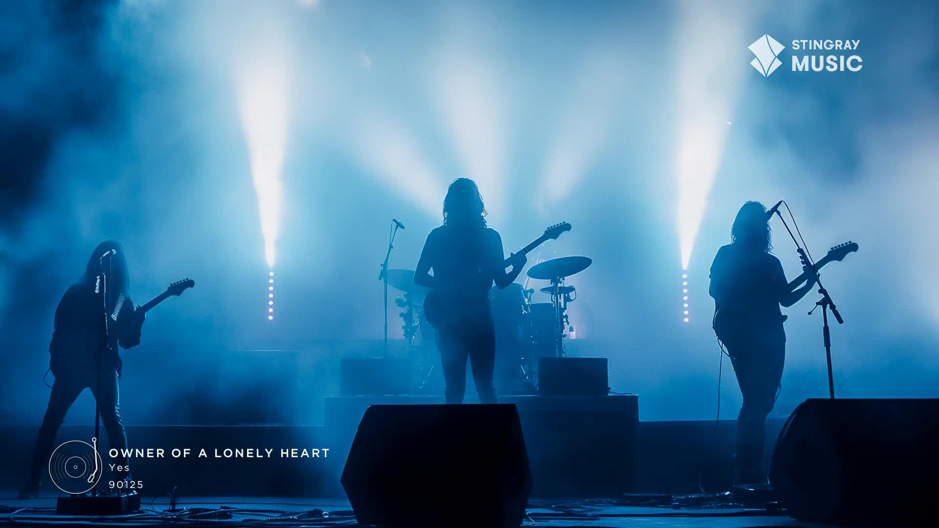 The band, silhouetted by stage lights, is playing "Owner of a Lonely Heart" on Stingray Classic Rock. The guitarist on the left is leaning into the mic, while the other two stand in the blue haze, guitars held high.
