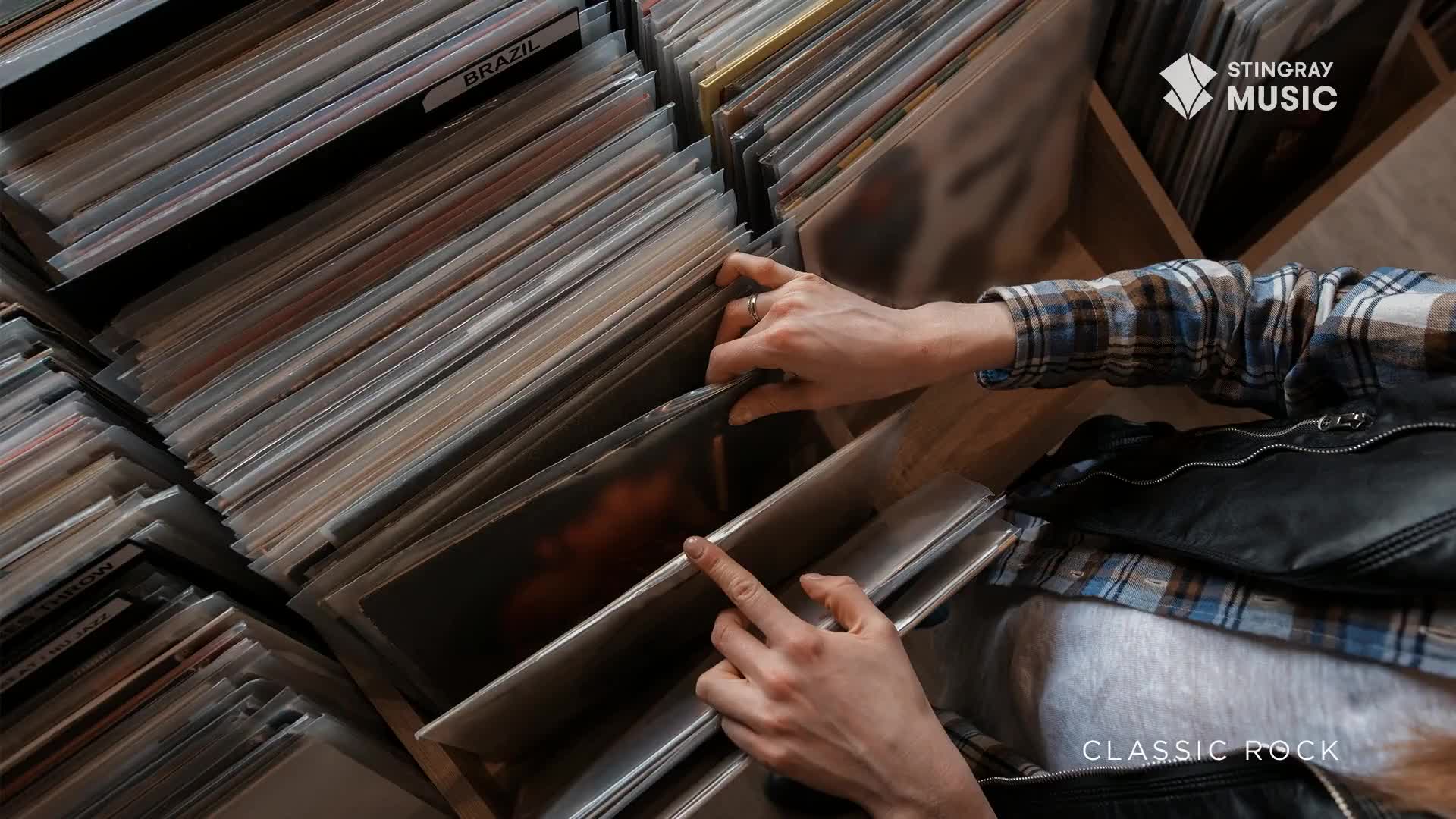 Someone's fingers are carefully sifting through a stack of vinyl records, likely hunting for a classic rock gem. The Stingray Music logo is visible, and the scene feels like a cozy record store in Canada.

