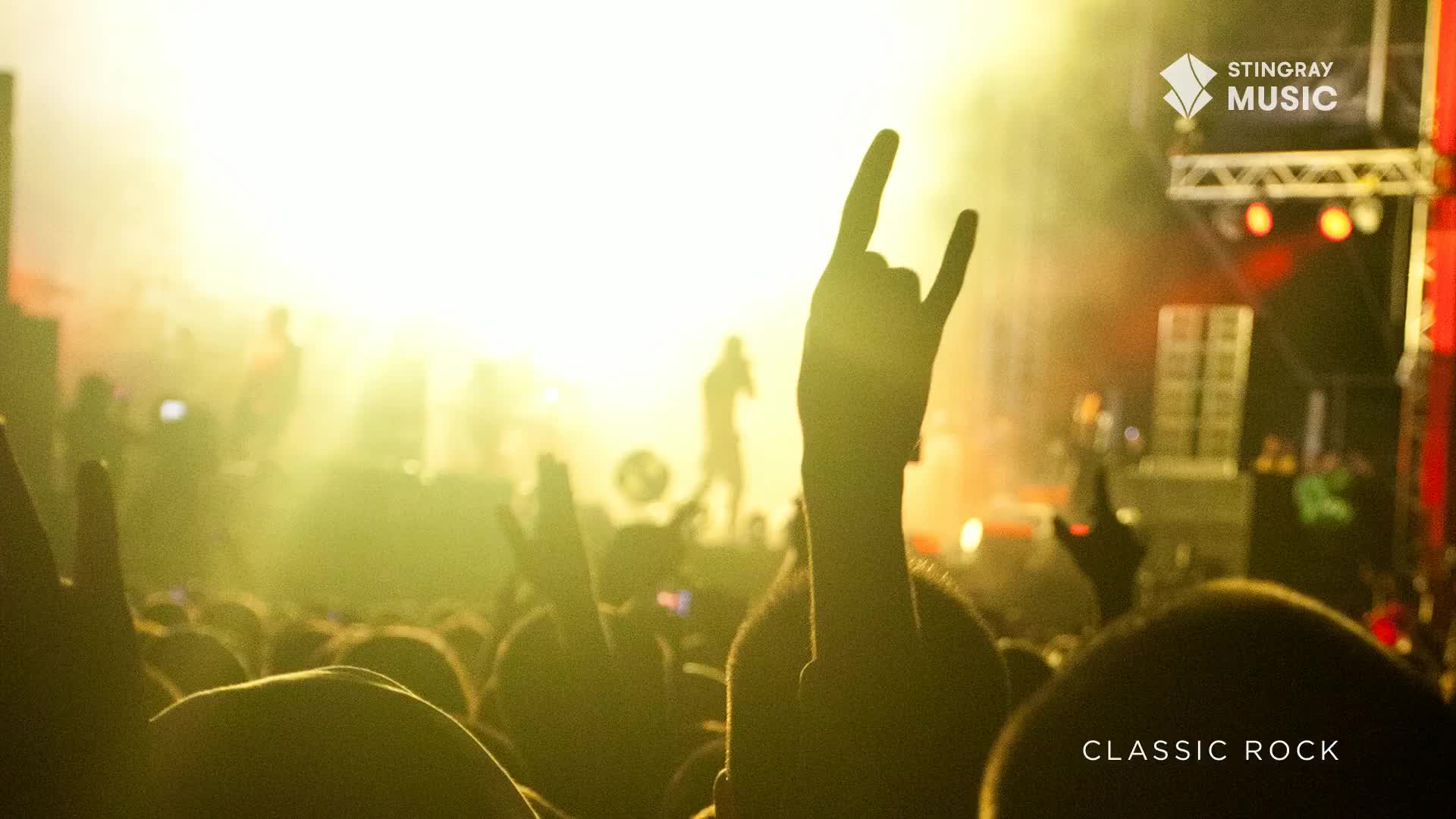 The crowd is a sea of raised hands, many forming the classic rock salute as the band plays on stage. A bright, hazy light washes over everything, making the silhouettes of the performers and the audience stand out.

