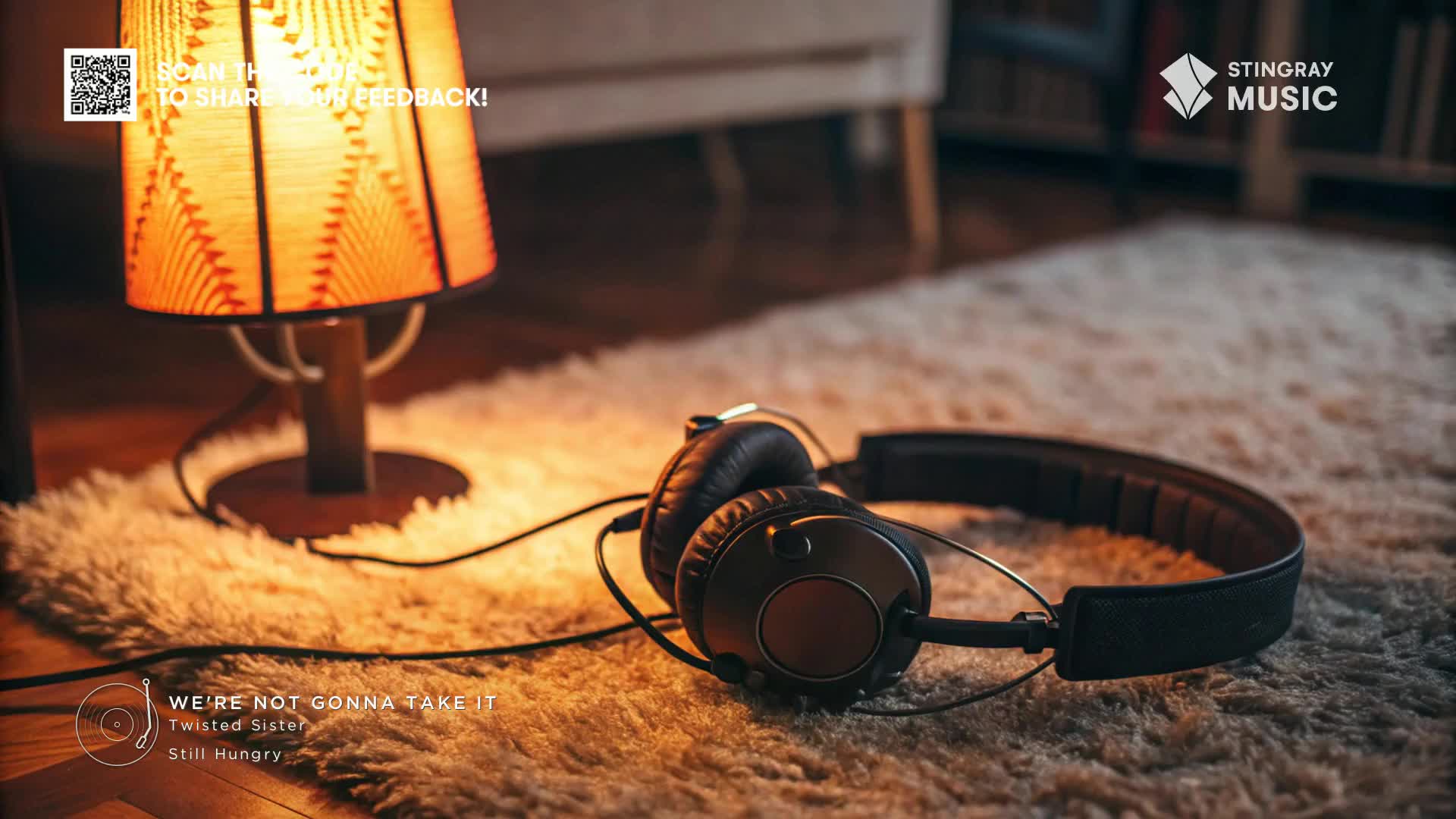 A warm lamp casts a soft glow on a fluffy rug, next to a pair of headphones. The Stingray Music logo is visible, and the song "We're Not Gonna Take It" is playing.
