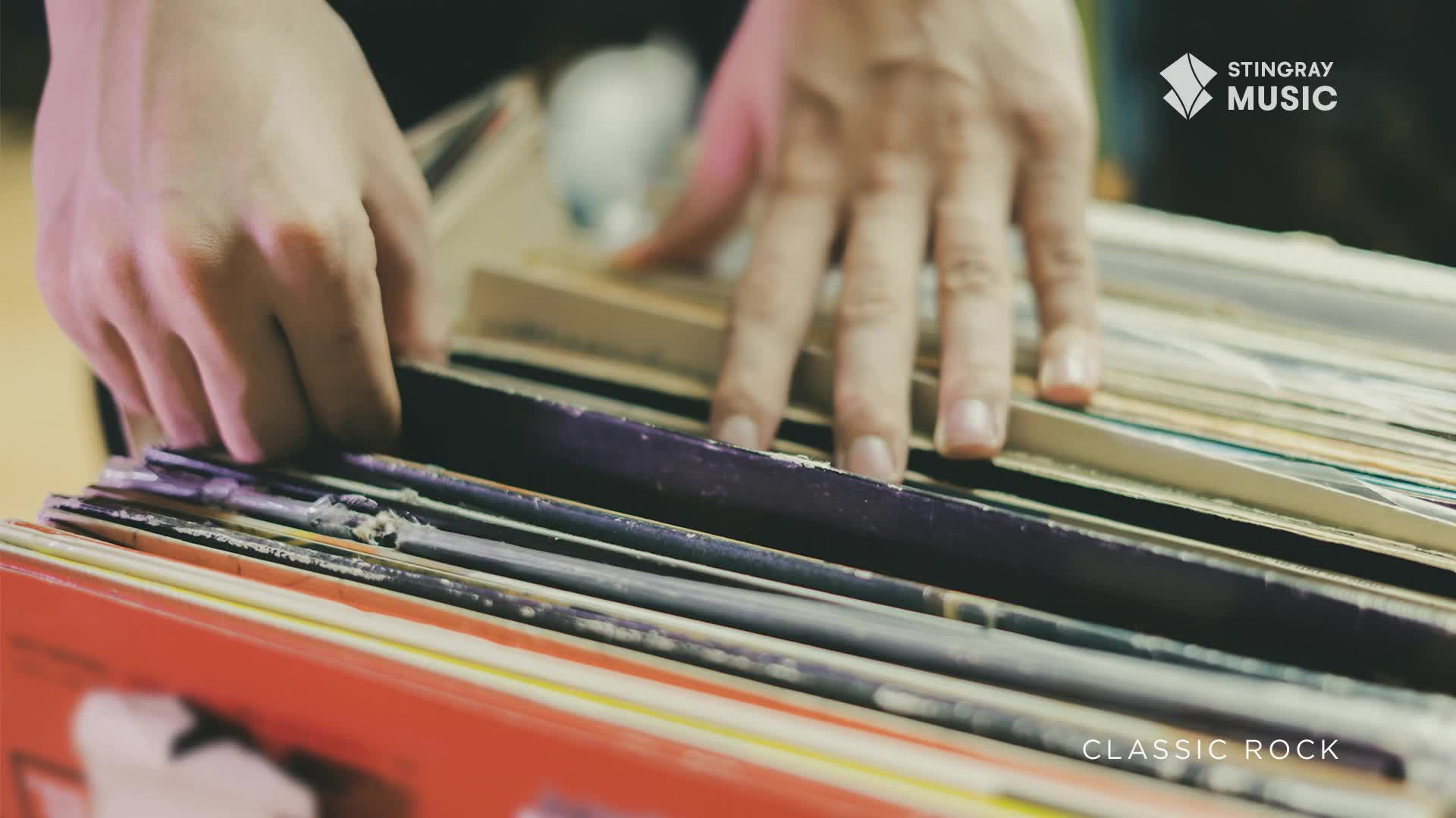 Fingers sift through a stack of vinyl records, searching for something. The collection, likely curated for Stingray Classic Rock, is neatly arranged in a red crate.
