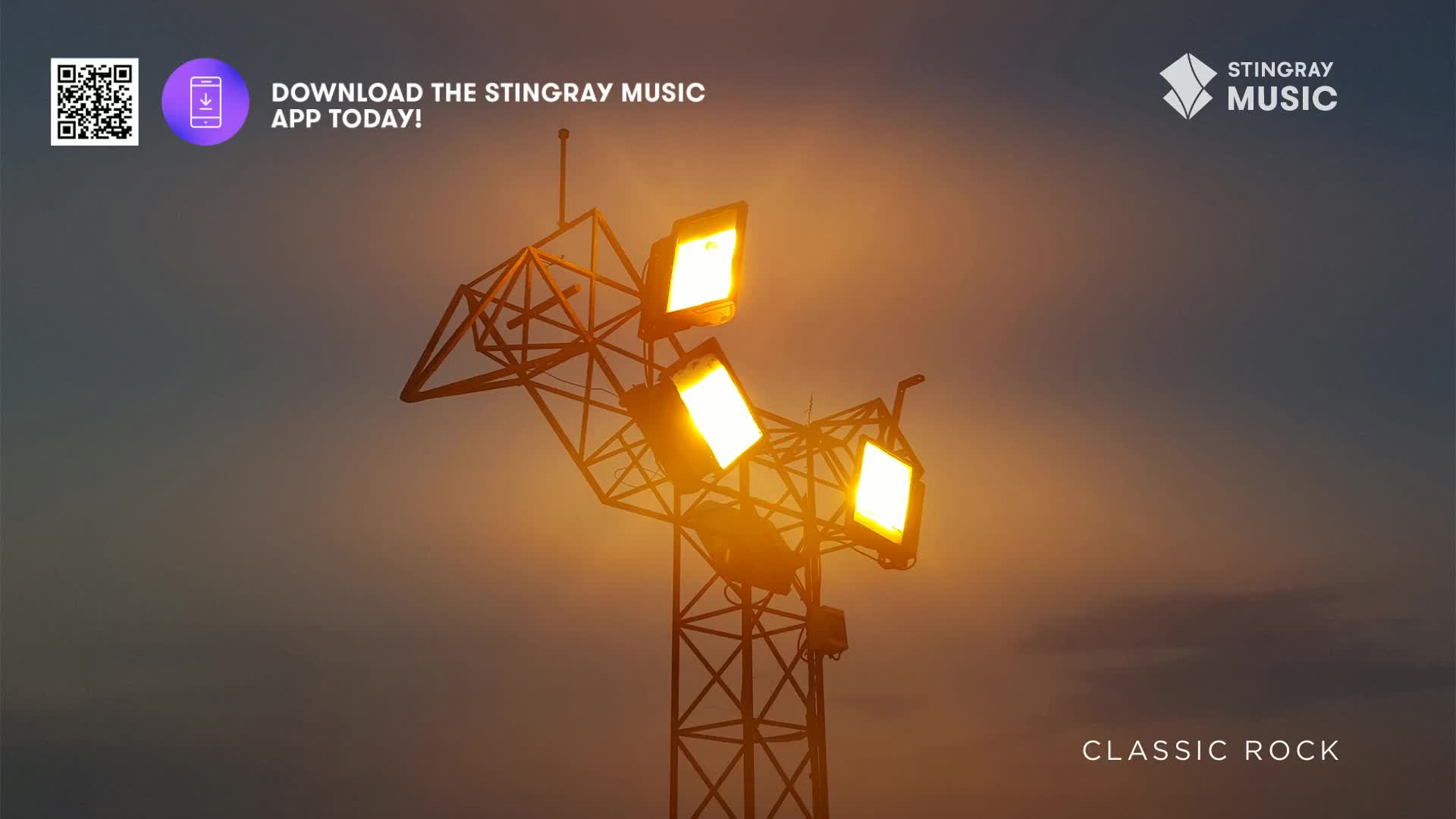 Above, a metal structure holds bright, square lights, illuminating the sky with a warm glow. The Stingray Classic Rock logo is visible, and the download app prompt is there too.
