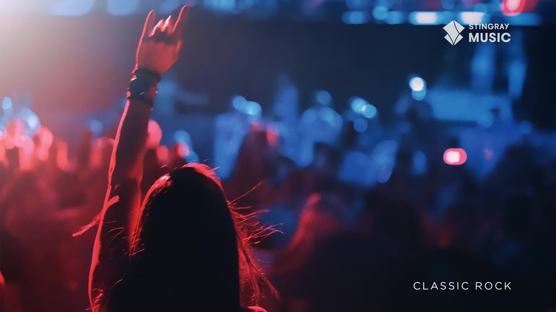 A hand, lit by red stage lights, shoots up in a rock and roll salute. The crowd is a sea of blurred shapes, bathed in blue and red hues, likely enjoying some classic rock from Stingray Music in Canada.
