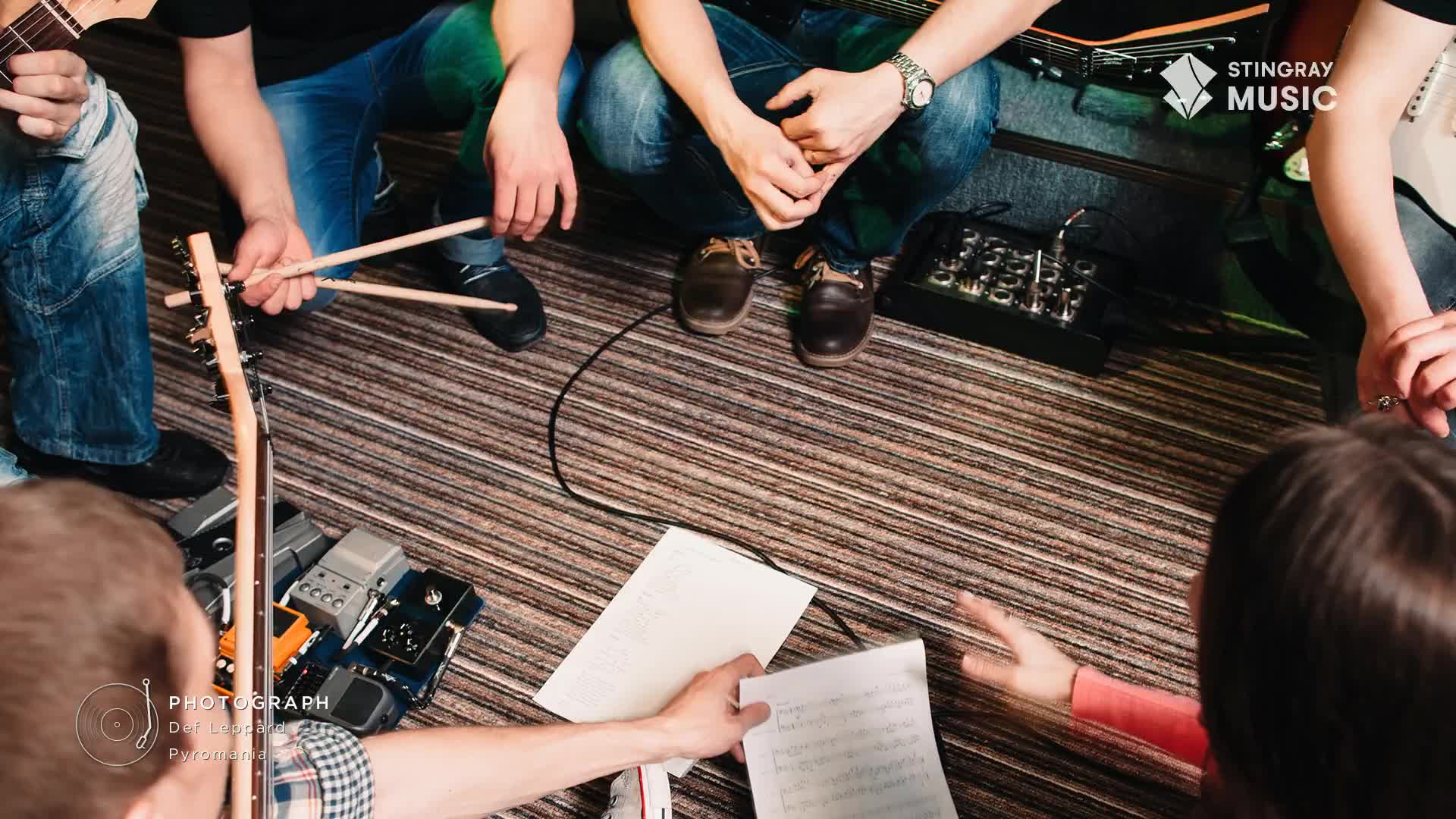 A group of musicians huddle on the floor, sheet music and instruments scattered around them. A pair of drumsticks rests in the hands of one person, ready to play.
