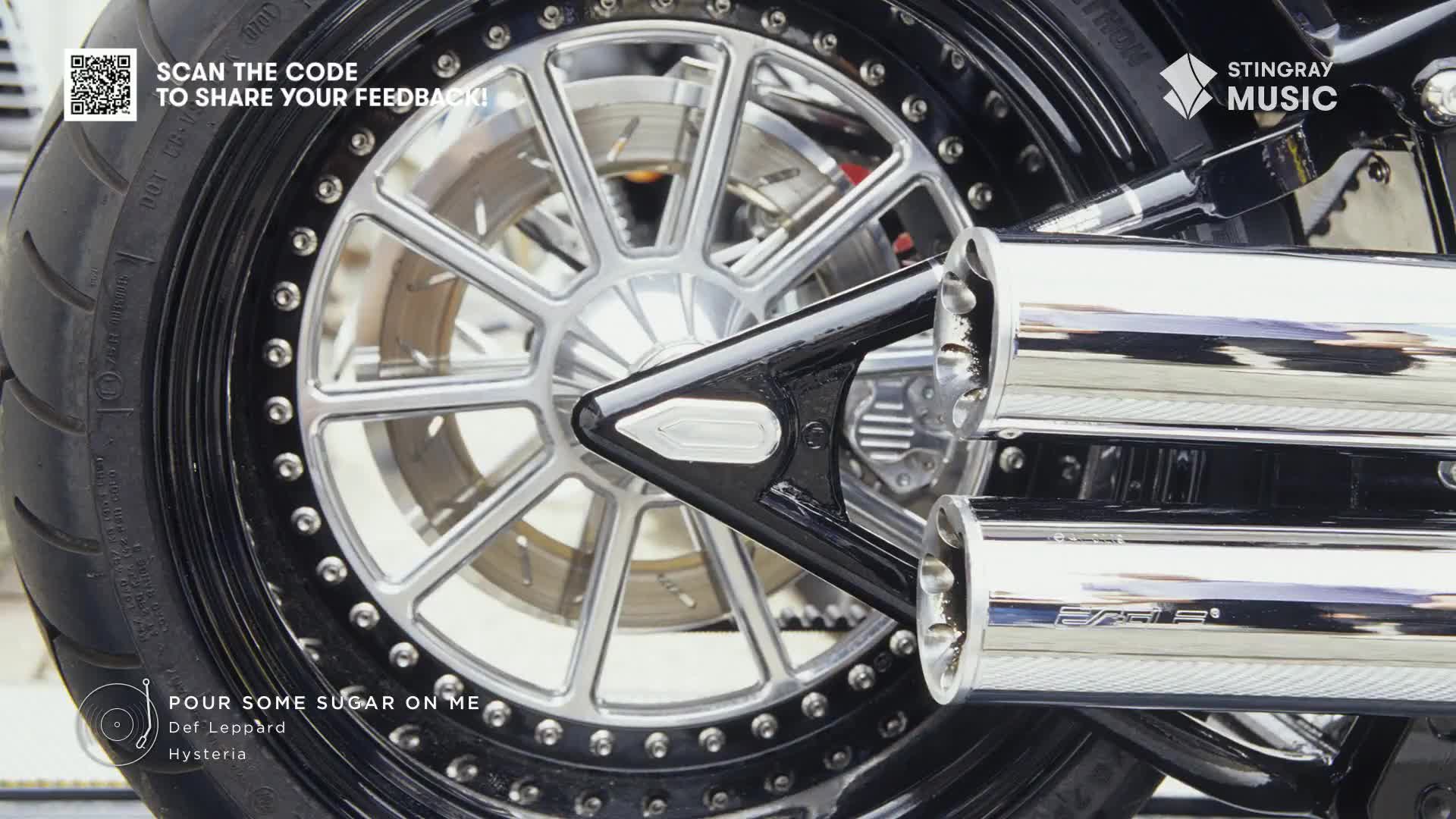The polished chrome exhaust pipes gleam, reflecting the light as Def Leppard's "Pour Some Sugar On Me" plays on Stingray Classic Rock. The black tire and silver wheel of a motorcycle are the focus, and the scene feels like it could be anywhere in Canada.
