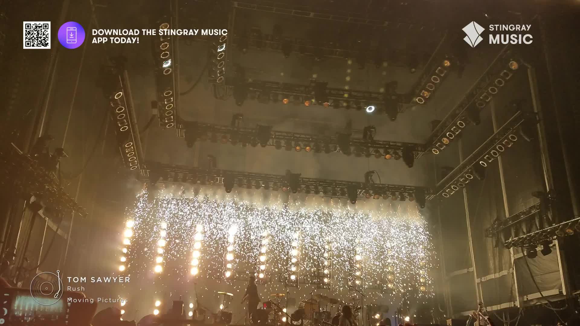 Sparks erupt from the stage as the band plays "Tom Sawyer." Above, the stage lights are aimed down, illuminating the performance for the crowd.
